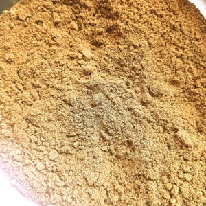 Könnte beinhalten: Nahaufnahme eines Haufens fein gemahlenen, hellbraunen Pulvers. Das Pulver hat ein strukturiertes Aussehen mit kleinen Granulaten und einer leicht unebenen Oberfläche. Die Farbe ist ein warmer, erdiger Ton, der auf ein Gewürz oder Kraut hindeutet.