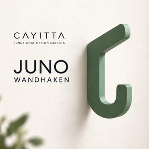 Könnte beinhalten: Ein salbeigrüner Wandhaken mit geometrischer, kantiger Form. Der Text CAYITTA FUNCTIONAL DESIGN OBJECTS und JUNO WANDHAKEN ist links neben dem Haken aufgedruckt. Der Haken hat eine flache, glatte Oberfläche und einen gebogenen unteren Bereich.