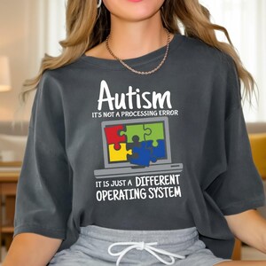 May include: Dark gray t-shirt with the word "Autism" and the phrase "It's not a processing error" printed in white. Below is a laptop graphic with puzzle pieces. The text "It is just a different operating system" is printed below the laptop graphic.
