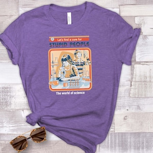 May include: Purple t-shirt with a graphic that reads "Let's find a cure for STUPID PEOPLE" and "The world of science." The graphic depicts two people in a lab. The shirt is on a light wood surface with sunglasses and plants.