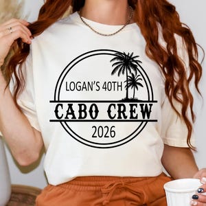 May include: Cream-colored t-shirt with a black circular design that reads "Logan's 40th Cabo Crew 2026" with palm tree silhouettes. The person is wearing orange shorts and holding a white mug.