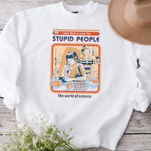May include: White crew neck sweatshirt with a retro graphic featuring two figures in a lab setting, the text "Let's find a cure for STUPID PEOPLE" and "The world of science". A brown hat rests on the sweatshirt.