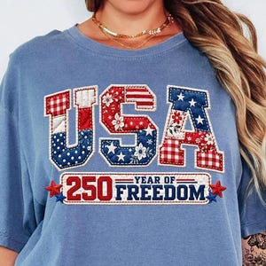 May include: A blue t-shirt with the word "USA" in a patchwork design featuring red, white, and blue patterns. Below the word "USA" is the text "250 YEAR OF FREEDOM" in a red and white design. The shirt has short sleeves.