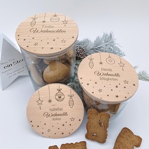 May include: Three glass jars with wooden lids, filled with cookies. The lids are engraved with festive designs and German text. One lid reads "Frohe Weihnachten," another "Davids Weihnachts Süßigkeiten," and the third "Isabellas Weihnachts Kekse."