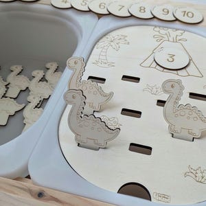 Könnte beinhalten: Holzspielzeug mit einem weißen Tablett, das Holzdinosaurierformen enthält. Das Tablett trägt die Nummer 3 und ein Bild eines Vulkans. Auf der Oberseite des Spielzeugs befinden sich auch Holzziffern von 4 bis 10.