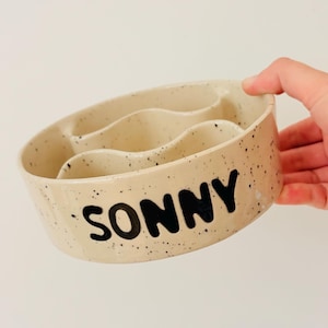 May include: A round, beige ceramic slow feeder bowl with a speckled finish and the name SONNY printed in bold black letters on the side. The interior features a wavy, raised divider structure designed to slow down the pace of eating.