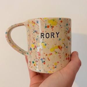 May include: A beige ceramic mug with colourful paint splatters in yellow, red, blue, and pink. The word "RORY" is printed in black on the front. The mug has a curved handle.