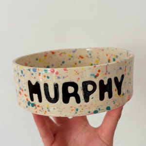 May include: A ceramic pet bowl with a cream base, decorated with colourful paint splatters. The name "MURPHY" is boldly displayed in black letters around the bowl's exterior. This bowl is designed for pets.