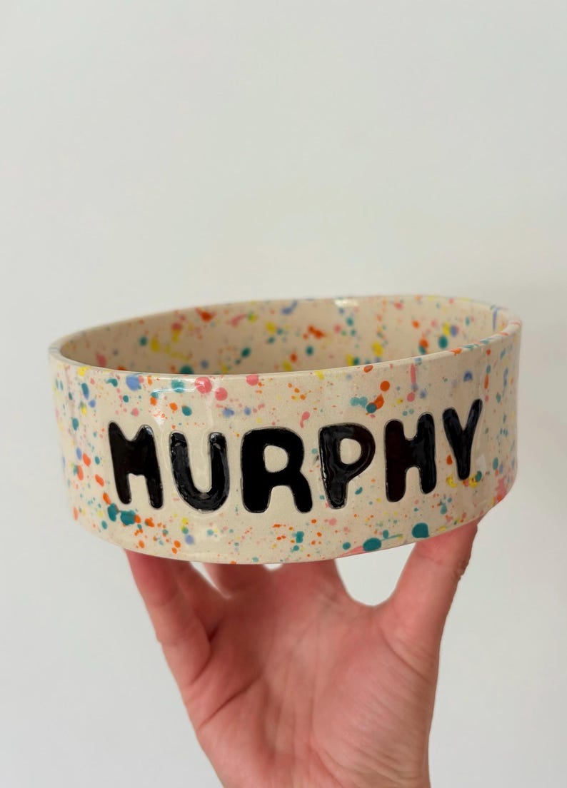 May include: A round ceramic bowl with a cream-colored base featuring a multi-colored paint splatter pattern in shades of blue, orange, pink, and green. The name MURPHY is written in large, bold, black capital letters on the front of the bowl.