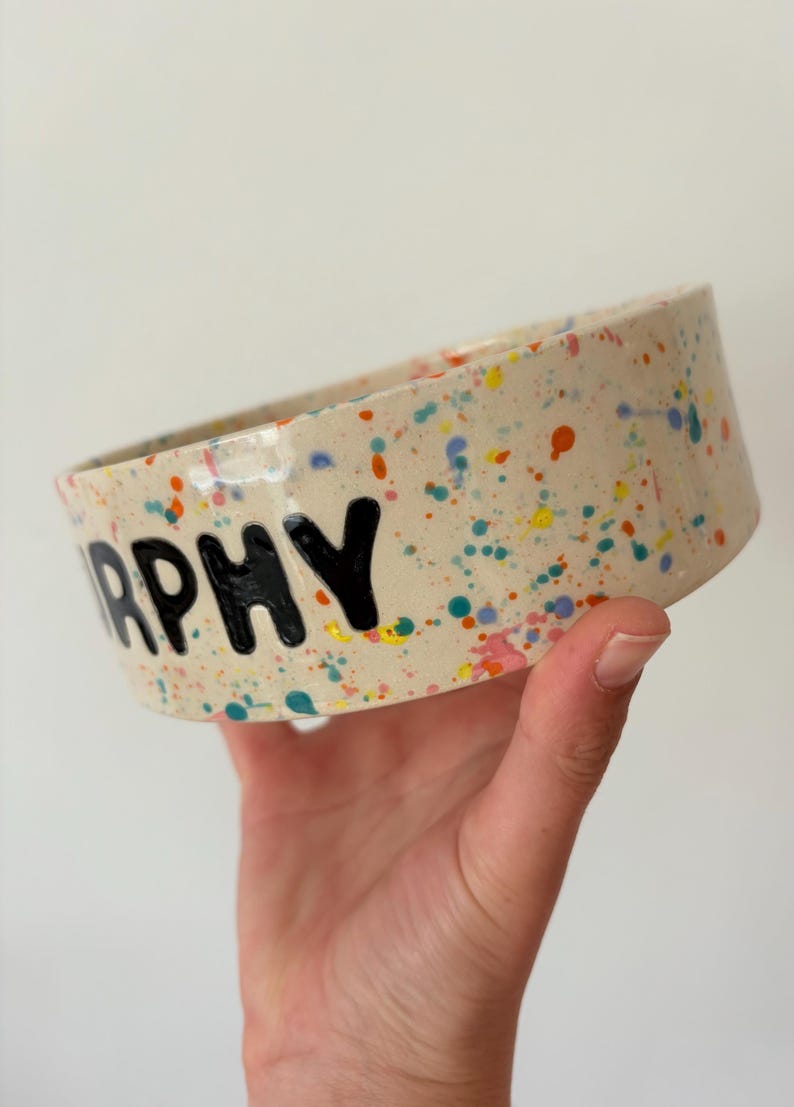 May include: A cylindrical ceramic bowl with a cream-colored base featuring a multi-colored paint splatter pattern in shades of blue, orange, pink, and yellow. The black text "RPHY" is clearly printed on the side of the bowl.