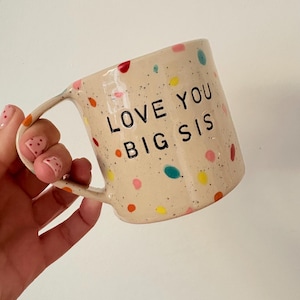 May include: A beige ceramic mug with colourful polka dots and the text "LOVE YOU BIG SIS" in black. The mug has a curved handle and is held at an angle.