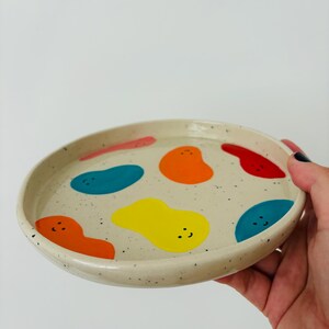 May include: A round, speckled ceramic plate with a cream-colored base. The plate is decorated with colorful, abstract shapes in red, orange, yellow, and blue. The plate is held by a hand against a white background.