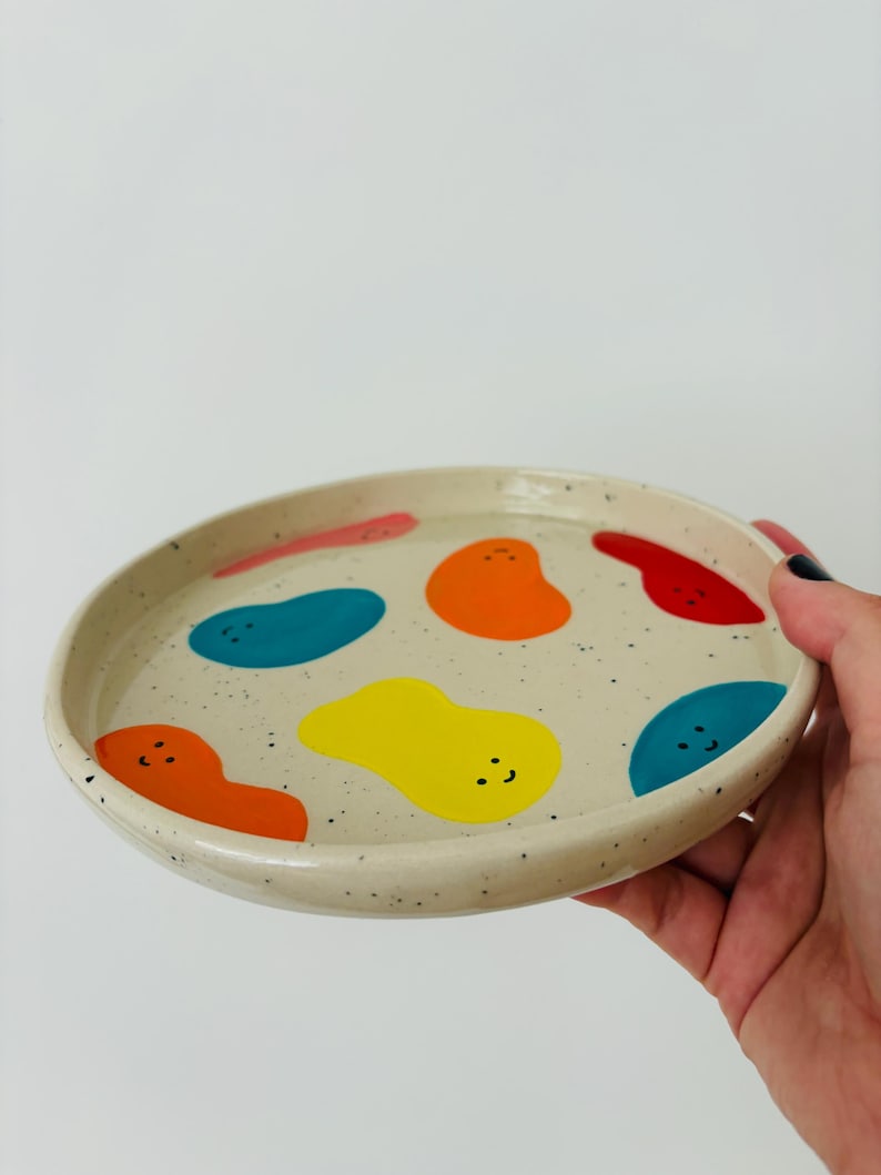 May include: A round, speckled ceramic plate with a cream-colored base. The plate is decorated with colorful, abstract shapes in red, orange, yellow, and blue. The plate is held by a hand against a white background.