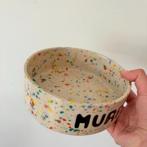 May include: A round ceramic bowl with a cream-colored base featuring a multi-colored paint splatter pattern in shades of blue, yellow, orange, and pink. The exterior of the bowl displays the black hand-painted text MURP clearly visible.