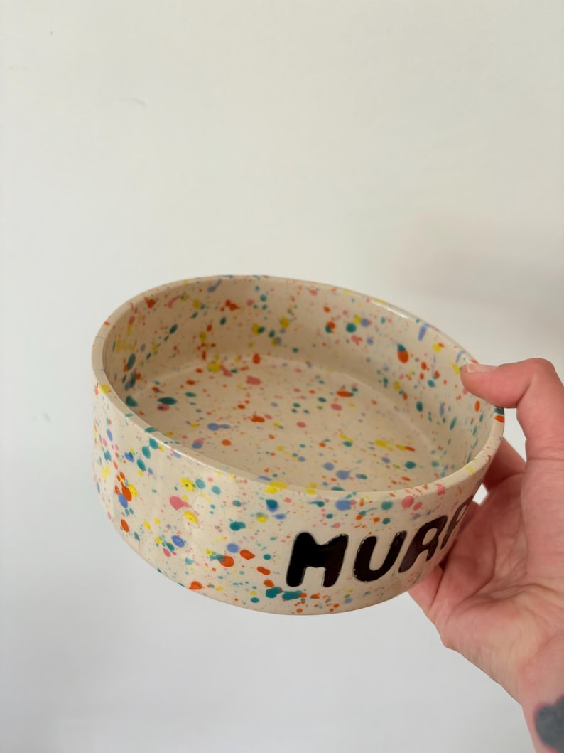 May include: A round ceramic bowl with a cream-colored base featuring a multi-colored paint splatter pattern in shades of blue, yellow, orange, and pink. The exterior of the bowl displays the black hand-painted text MURP clearly visible.