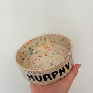 May include: A round ceramic bowl with a cream-colored base featuring a multi-colored paint splatter pattern in shades of orange, blue, and yellow. The exterior of the bowl has the name MURPHY printed in bold, black, hand-painted capital letters.