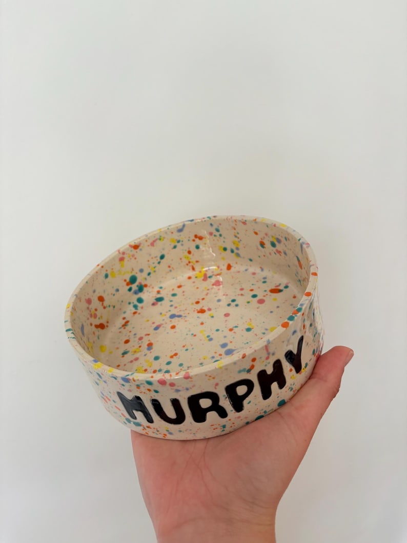 May include: A round ceramic bowl with a cream-colored base featuring a multi-colored paint splatter pattern in shades of orange, blue, and yellow. The exterior of the bowl has the name MURPHY printed in bold, black, hand-painted capital letters.