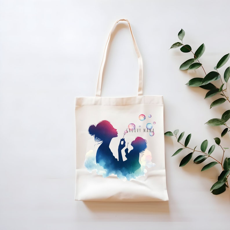 Op de afbeelding: Een witte canvas tote bag met een kleurrijk silhouet van een moeder en kind die zeepbellen blazen. De tekst "Groovy Mama" is op de tas gedrukt.