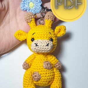 May include: A yellow crochet giraffe keyring with brown detailing and a blue flower charm. The giraffe has black eyes and a beige muzzle. The keyring includes a metal ring and the word "PDF" in a yellow circle.
