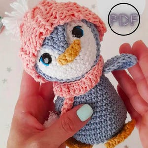 Könnte beinhalten: Ein gehäkelter Pinguin, der eine rosa Mütze und einen Schal trägt. Der Pinguin ist grau mit einem weißen Bauch und einem gelben Schnabel. Die Mütze hat einen weißen Bommel oben drauf.