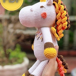 May include: A white crocheted unicorn doll with a yellow horn, mane, and hooves. The unicorn has a small orange heart on its cheek and a colorful tail. The doll is held in a person's hand.