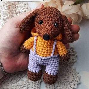 Può includere: Un bassotto di uncinetto marrone che indossa una camicia gialla con una salopette viola. Il bassotto tiene un piccolo cuore bianco nelle sue zampe.