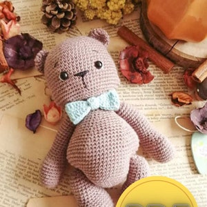 Puede incluir: Un oso de peluche de ganchillo hecho a mano en color malva suave, con una pajarita azul claro. El oso tiene ojos de botón negros y una pequeña nariz negra. La imagen incluye flores secas y un logotipo PDF.