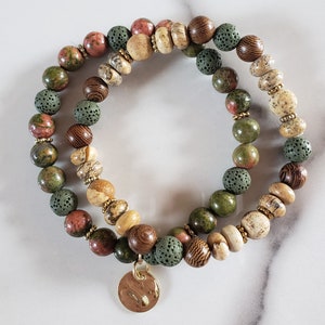 May include: A set of two bracelets featuring green, brown, and pink beads. The bracelets are made of various materials, including wood, lava rock, and gemstone. The bracelets are accented with gold spacers and a gold charm.