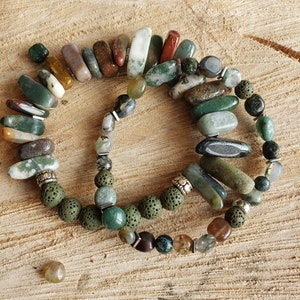 May include: Two bracelets made with green, brown, and gray agate stones, lava rock beads, and silver spacers. The bracelets are on a wooden surface.