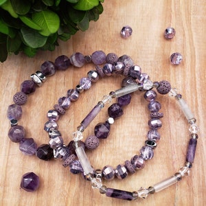 May include: Three amethyst beaded bracelets with various bead shapes and sizes. The bracelets feature faceted, round, and cylindrical beads in shades of purple and silver. Some beads have a textured, porous appearance. A loose amethyst crystal and several loose beads are scattered nearby.