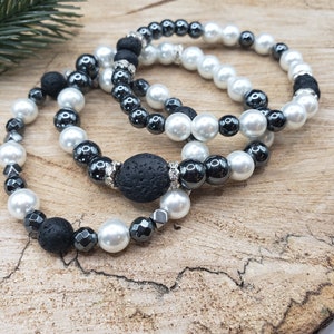 May include: Three beaded bracelets featuring a mix of white faux pearls, black lava stones, and hematite beads. The bracelets are arranged on a rustic wooden surface, with a sprig of greenery in the background. Each bracelet has a different design.