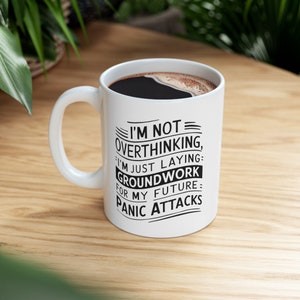 Puede incluir: Taza de café de cerámica blanca con asa negra. La taza tiene texto negro que dice "No estoy pensando demasiado, solo estoy sentando las bases para mis futuros ataques de pánico."