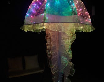 May include: A large, illuminated jellyfish-shaped lamp with a translucent, ruffled skirt and a dome-shaped top. The lamp is lit with colorful LED lights, creating a vibrant display of pink, blue, green, and yellow hues. The lamp hangs from the ceiling in a dark room.