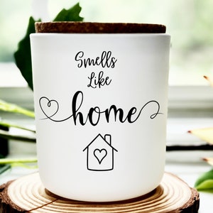Puede incluir: Un tarro de vela blanco con tapa de corcho y el texto "Smells Like Home" en letra negra. Un pequeño icono de casa con un corazón dentro está debajo del texto.