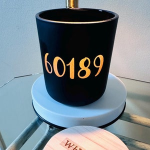May include: A black candle holder with the number "60189" in gold lettering. The candle holder sits on a white base, and a wooden lid with the word "WHEATON" is next to it. The candle holder is on a glass table.