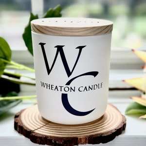 May include: White candle with a wooden lid and a black and white logo that says "Wheaton Candle" with a stylised "W" and "C". The candle is sitting on a wooden slice.