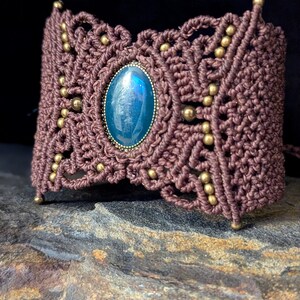 Könnte beinhalten: Handgefertigtes Makramee-Armband in Braun mit einem ovalen, blau schimmernden Edelstein in der Mitte. Das Armband ist mit kleinen, goldfarbenen Perlen verziert und hat ein breites Manschettendesign. Dunkler Hintergrund.