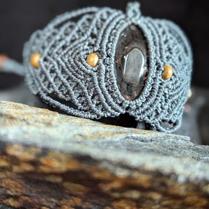 Könnte beinhalten: Ein graues Makramee-Armband mit einem dunkelgrauen Edelstein und goldenen Perlen.