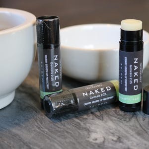 May include: Three black and green lip balm tubes with the text "NAKED Genesis 2:25 Creamy Smooth Lip Protectant" on the labels. The lip balms are on a gray surface.