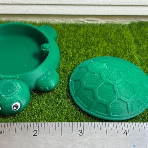 Puede incluir: Un arenero de pl&aacute;stico verde en forma de tortuga con una tapa separada. El arenero tiene un di&aacute;metro de unos 13 cm y la tapa de unos 15 cm de di&aacute;metro.