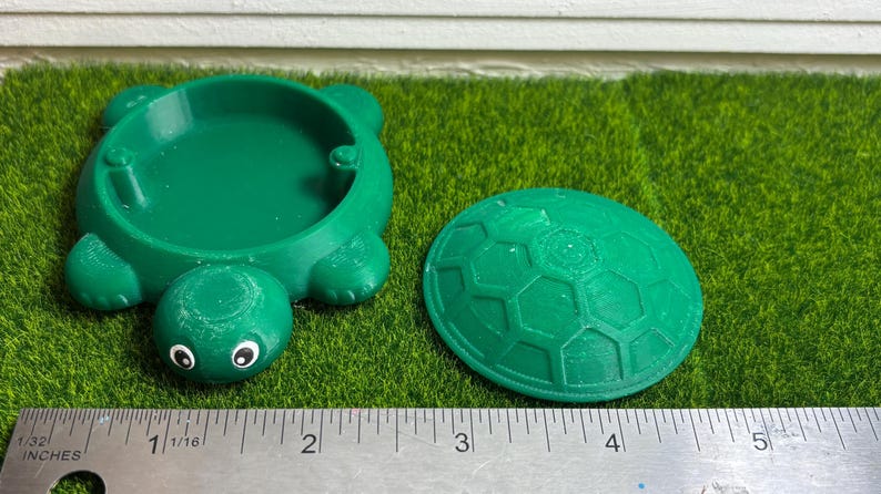 Puede incluir: Un arenero de pl&aacute;stico verde en forma de tortuga con una tapa separada. El arenero tiene un di&aacute;metro de unos 13 cm y la tapa de unos 15 cm de di&aacute;metro.