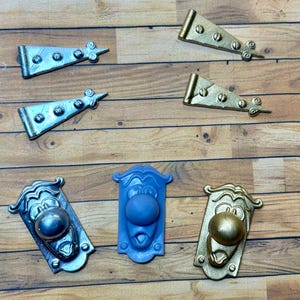 Puede incluir: Surtido de herrajes decorativos, que incluyen bisagras y pomos plateados y dorados. Los pomos tienen forma de cara con un pomo redondo. El hardware está dispuesto sobre una superficie de madera.