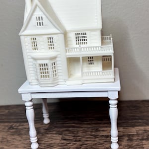 May include: A white miniature house with a detailed facade, including windows, a balcony, and a chimney, sits atop a small white table. The house has a classic architectural design. The table has four decorative legs.
