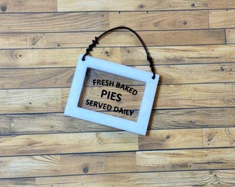 Puede incluir: Un letrero blanco con texto negro que dice "Fresh Baked Pies Served Daily". El letrero tiene un colgador de alambre negro.