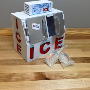 May include: A white ice house with a red "ICE" sign and a blue and white sign that reads "PACKAGED ICE CRYSTAL CLEAR". There are two bags of ice on the counter in front of the ice house.