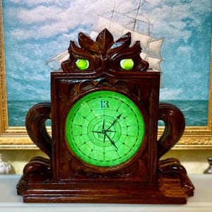 Pode incluir: Um relógio de madeira com um mostrador verde e o número 13. O relógio tem um design de bússola e está emoldurado em uma moldura marrom escura e ornamentada. A parte superior do relógio tem dois olhos verdes brilhantes.