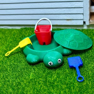 May include: A green turtle-shaped sandbox with a red bucket and two plastic shovels, one yellow and one blue. The sandbox is on a green artificial grass surface.