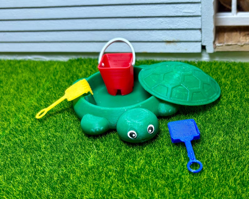Puede incluir: Un arenero con forma de tortuga verde con un cubo rojo y dos palas de pl&aacute;stico, una amarilla y una azul. El arenero est&aacute; sobre una superficie de c&eacute;sped artificial verde.