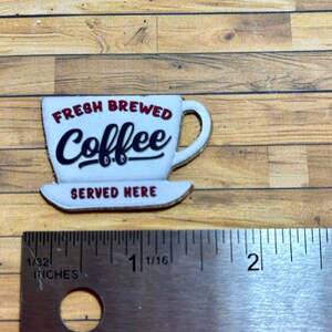 May include: White ceramic coffee cup with red lettering that reads "Fresh Brewed Coffee Served Here". The cup is on a wooden surface with a ruler for scale.
