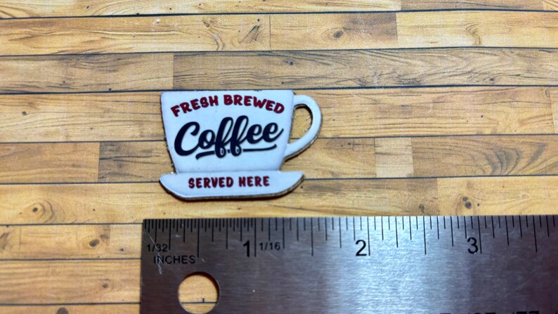 May include: White ceramic coffee cup with red lettering that reads "Fresh Brewed Coffee Served Here". The cup is on a wooden surface with a ruler for scale.